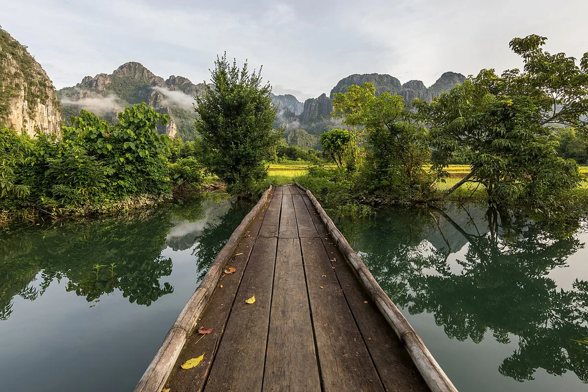 Front view of a wooden footbridge over a lagoon trees and mountains in Vang Vieng Laos — 1